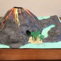 Maquette d'un volcan en éruption, avec de la lave rouge et orange dégoulinant sur ses flancs. Un paysage miniature avec une rivière, des palmiers et une petite île. Un projet pour fabriquer un volcan.