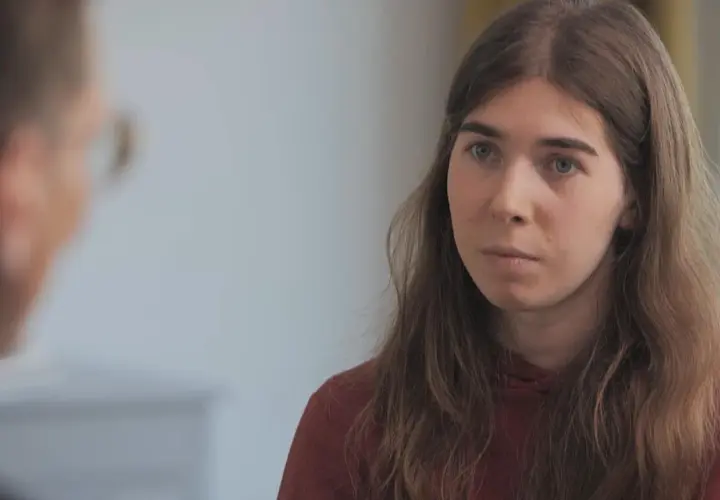 Une jeune femme aux cheveux longs et châtains, portant un sweat à capuche bordeaux, écoute attentivement une personne dont on ne voit que le dos. Elle semble réfléchir à son parcours pour changer de sexe.