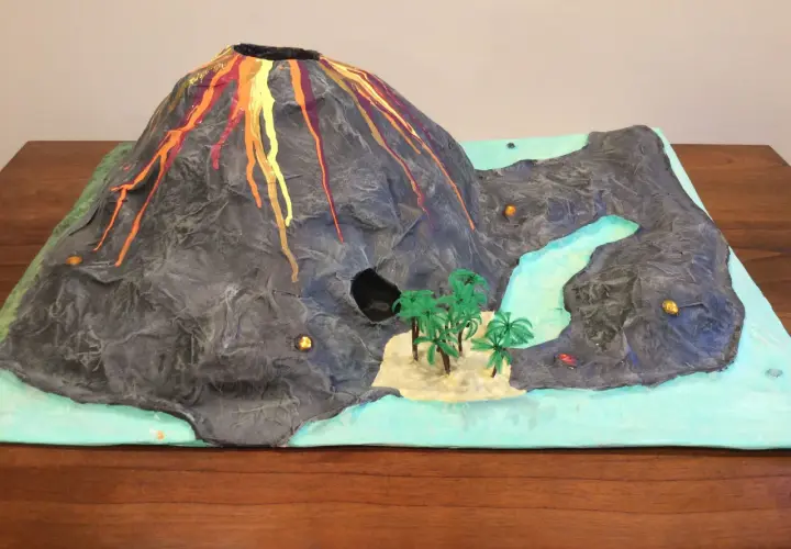 Maquette d'un volcan en éruption, avec de la lave rouge et orange dégoulinant sur ses flancs. Un paysage miniature avec une rivière, des palmiers et une petite île. Un projet pour fabriquer un volcan.