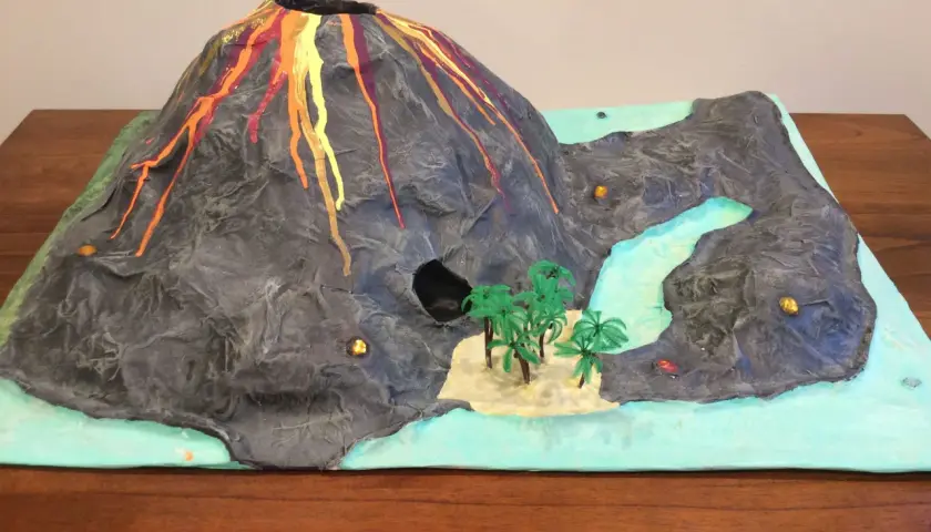 Maquette d'un volcan en éruption, avec de la lave rouge et orange dégoulinant sur ses flancs. Un paysage miniature avec une rivière, des palmiers et une petite île. Un projet pour fabriquer un volcan.