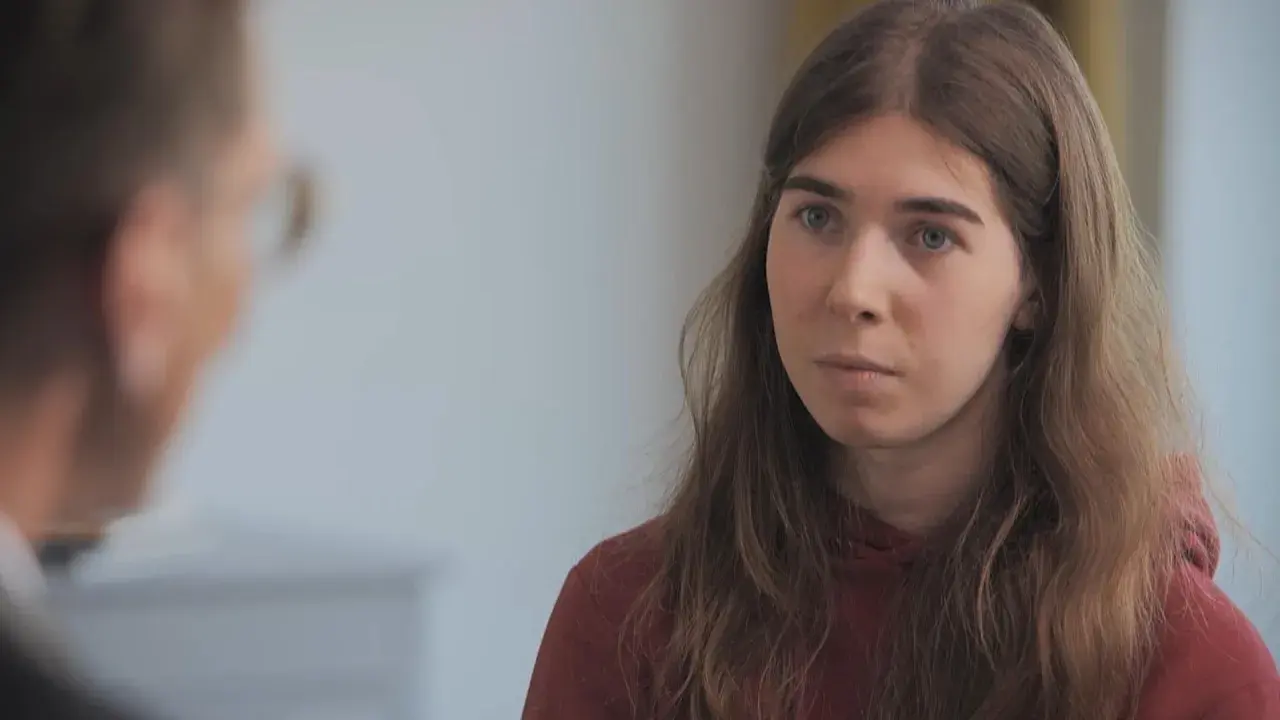 Une jeune femme aux cheveux longs et châtains, portant un sweat à capuche bordeaux, écoute attentivement une personne dont on ne voit que le dos. Elle semble réfléchir à son parcours pour changer de sexe.