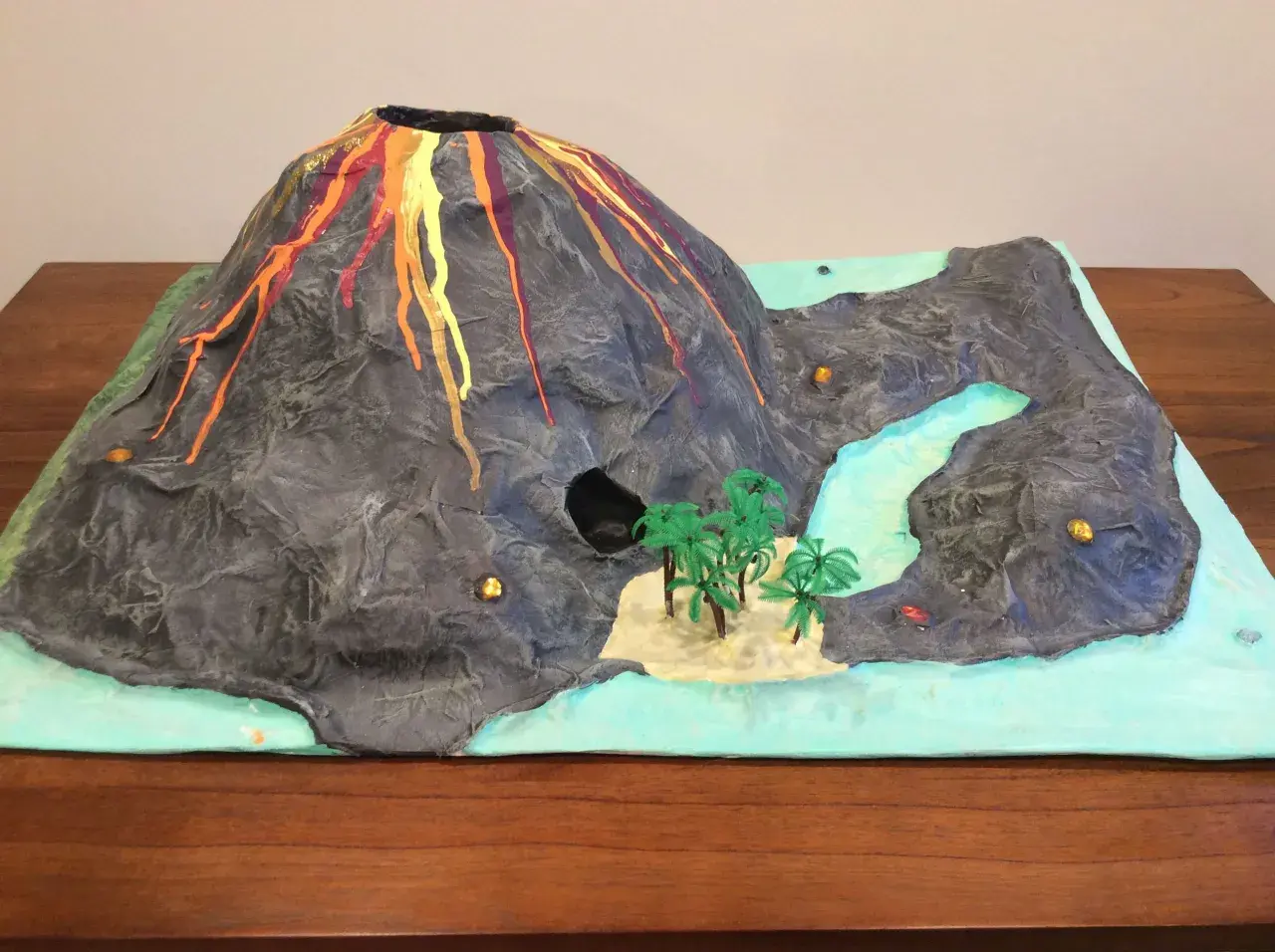 Maquette d'un volcan en éruption, avec de la lave rouge et orange dégoulinant sur ses flancs. Un paysage miniature avec une rivière, des palmiers et une petite île. Un projet pour fabriquer un volcan.