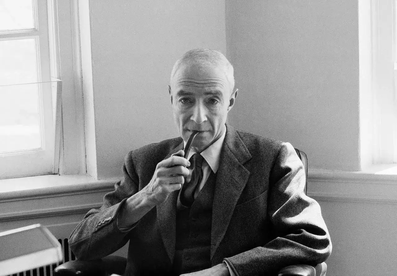 Robert Oppenheimer, le père de la bombe atomique, fume sa pipe, pensif, devant une fenêtre.