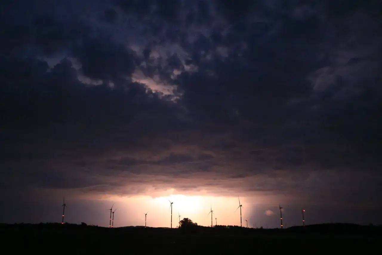 Un ciel orageux, des éoliennes illuminées par un éclair de chaleur, la nuit tombe sur le paysage.
