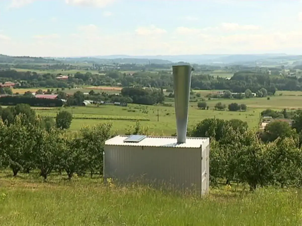 Un canon anti grêle se dresse sur une colline, veillant sur un paysage verdoyant de champs et de vergers sous un ciel bleu.