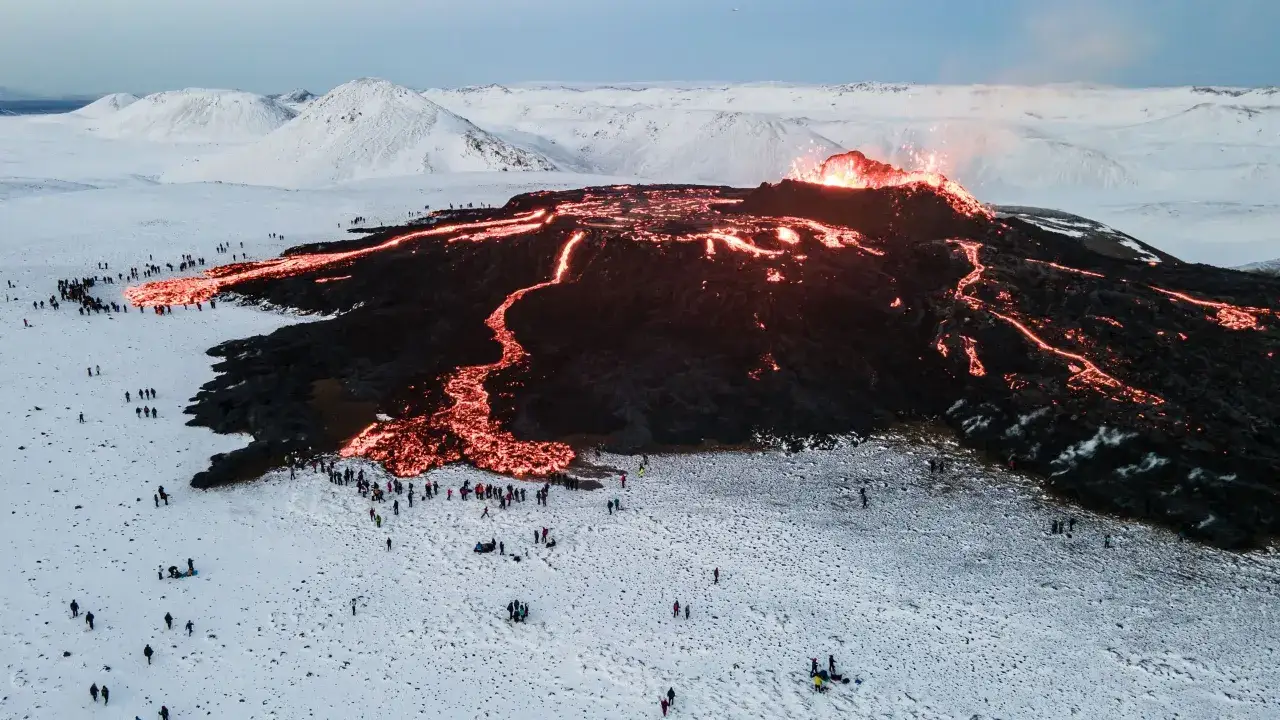 Éruption volcanique en Islande : la lave incandescente serpente sur le flanc d'un volcan, sous un ciel gris, tandis que des spectateurs observent ce spectacle impressionnant.