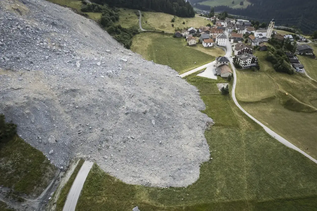 Vue aérienne d'un village suisse touché par un glissement de terrain. Des rochers et débris recouvrent une partie du paysage verdoyant et des habitations.